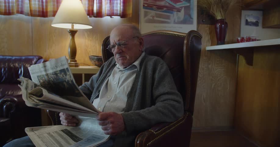 An old man sitting in a lounge chair focusing intently on reading a newspaper