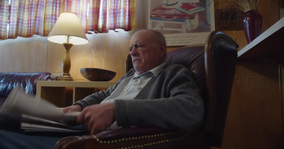 Elderly man reading a newspaper while sitting at home takes off his glasses