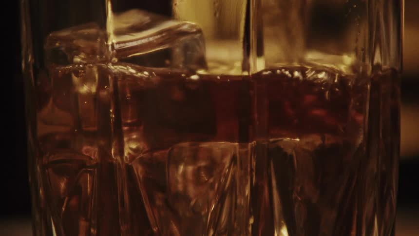 Glass with ice cubes fills up with liquor on a dimly lit room