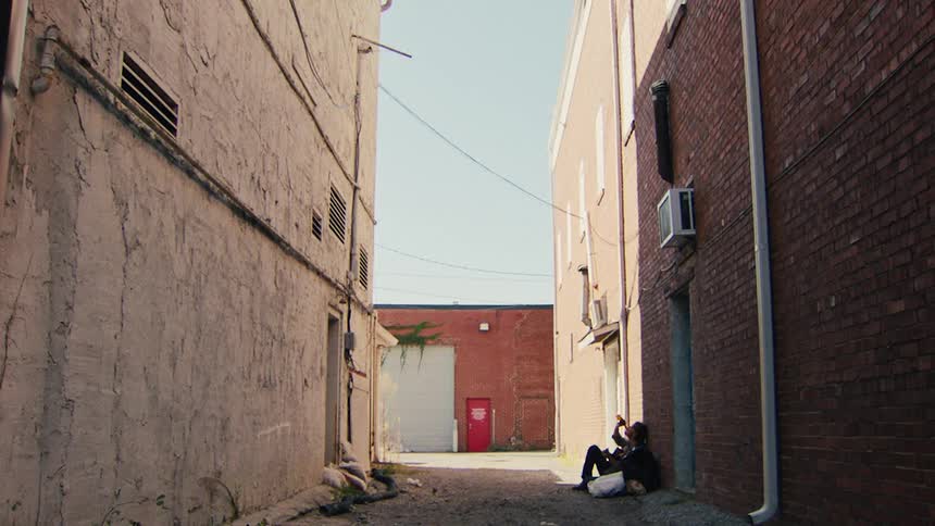 Man walks to end of alley and stands watching man sitting against wall and drinking