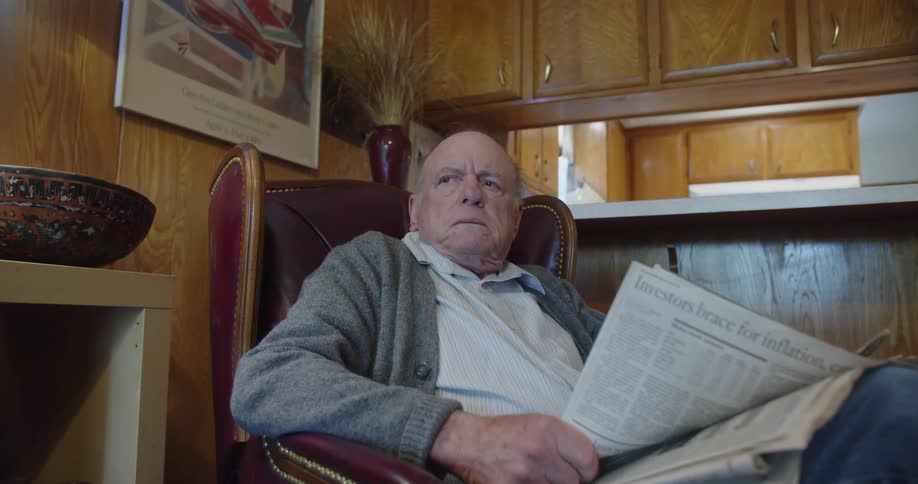A man sitting in a chair reading a newspaper takes off his glasses and grimaces in frustration