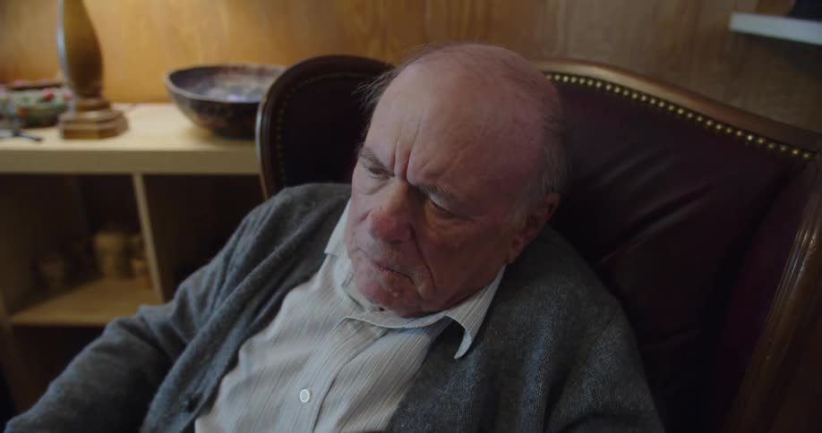 Elderly man sitting on a chair in the living room reading a newspaper takes his glasses off