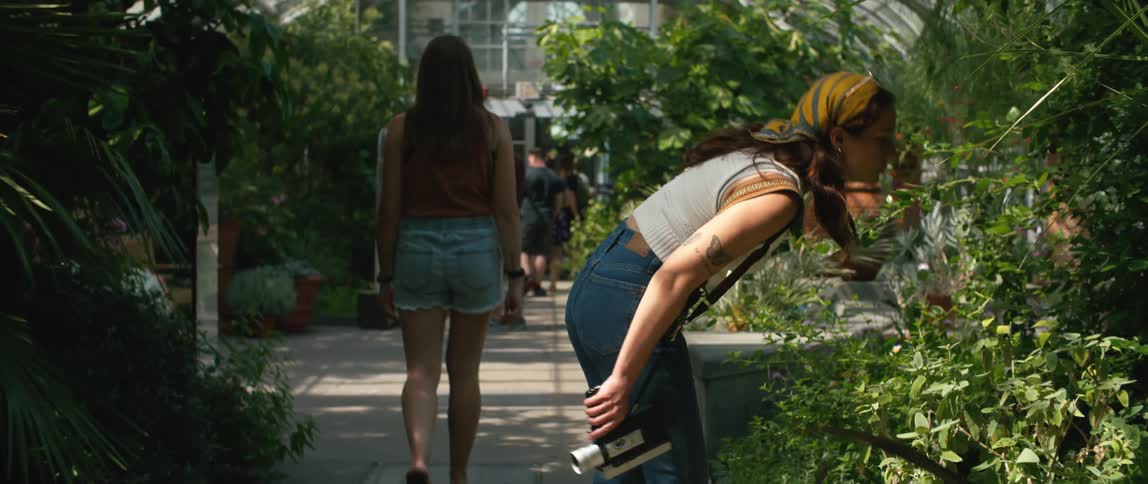 A group visits a green house and two teenage girls lag behind as one of them leans over to smell a plant before moving on