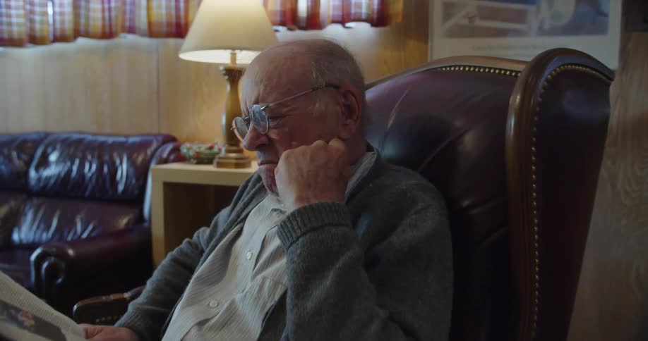 An old man sitting in a leather chair looks concerned as he reads a newspaper