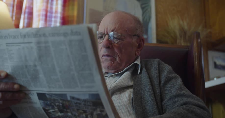 An elderly man sitting in a chair reading and holding a newspaper in his hands while grimacing and shaking his head
