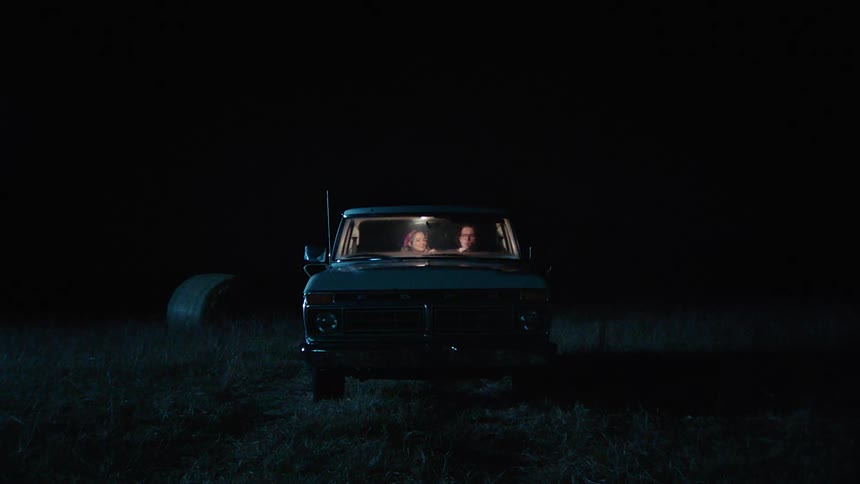 Man and woman sit in lit cab of truck parked in field at night and share smoke