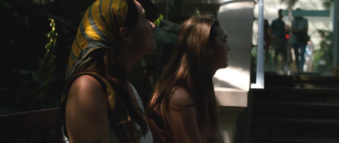 Two women are sitting outside on a bench looking around as people are coming out of a building
