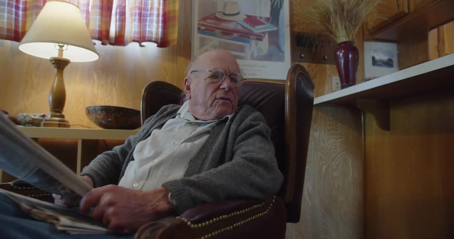 An old man in a lounge chair stops reading his newspaper to speak and engage with someone to his side