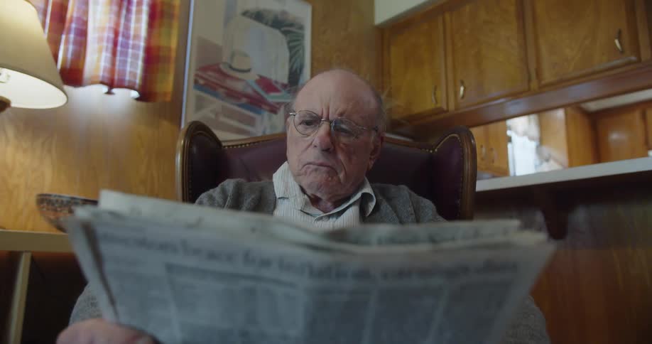 Elderly bald man with glasses gray cardigan and jeans reads newspaper shaking his head
