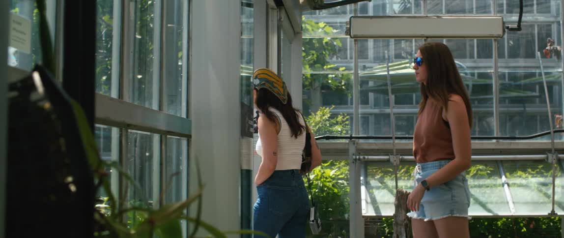 Two young women walking through a green house with windows on all sides as they walk outdoors