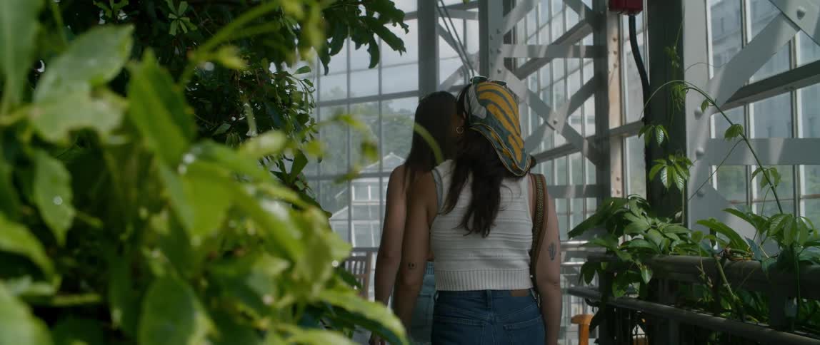 Two women are walking in a room filled with plants looking around as the sun shines in the windows