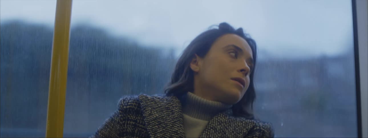 A worried looking woman sits on a bus with her head leaning back against the window and then looks up before leaning her head back again