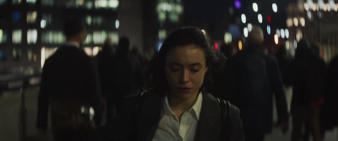 A worried looking woman walks down a busy city street at night and looks at her cell phone