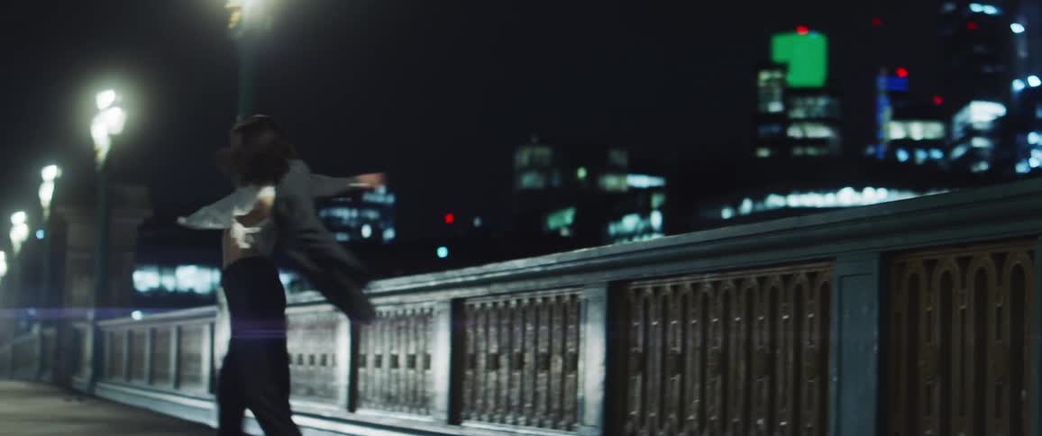 Female dancer with gray jacket white top and black pants dances on the bridge against the city skyline at night
