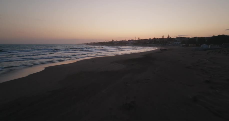 Waves roll onto uneven beach bordering town under red tinted sky