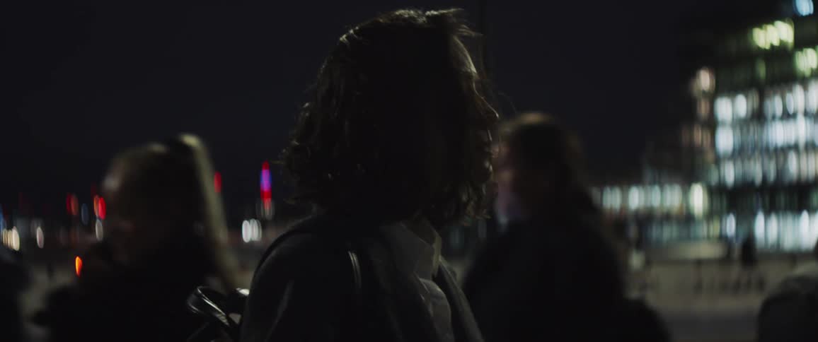 A close up of a woman standing outside at night on a sidewalk with her eyes closed as people walk by around her