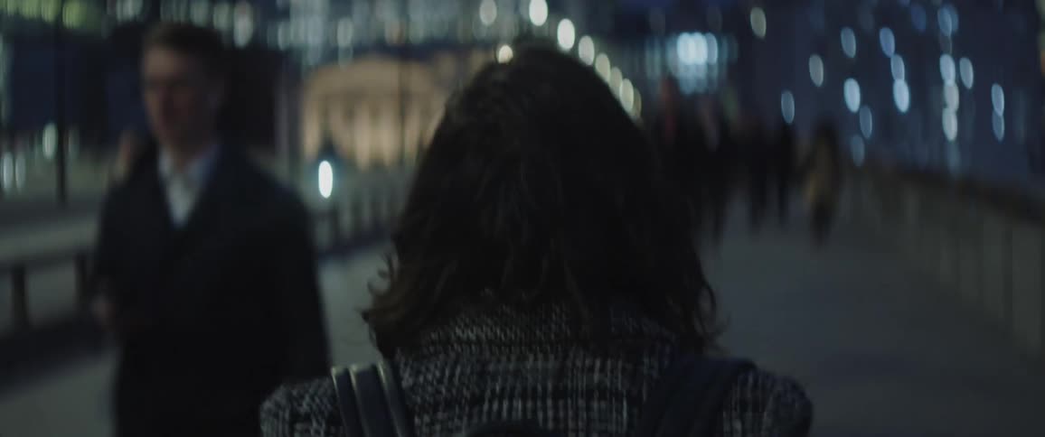 A woman walks hurriedly through a busy city bridge at night running her hand through her hair as she does it
