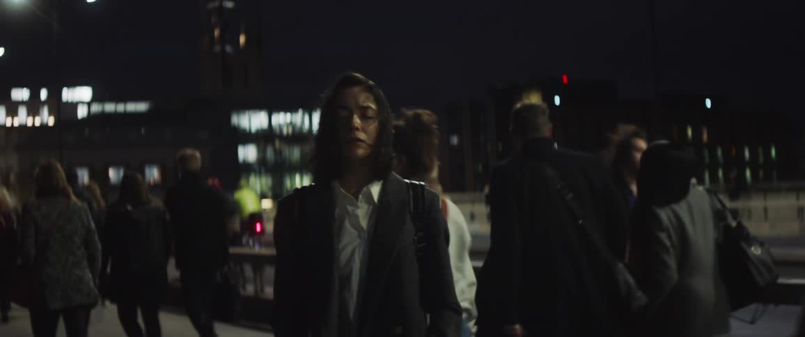 A woman is standing on a busy downtown sidewalk as many people walk by her as she stands with her eyes closed