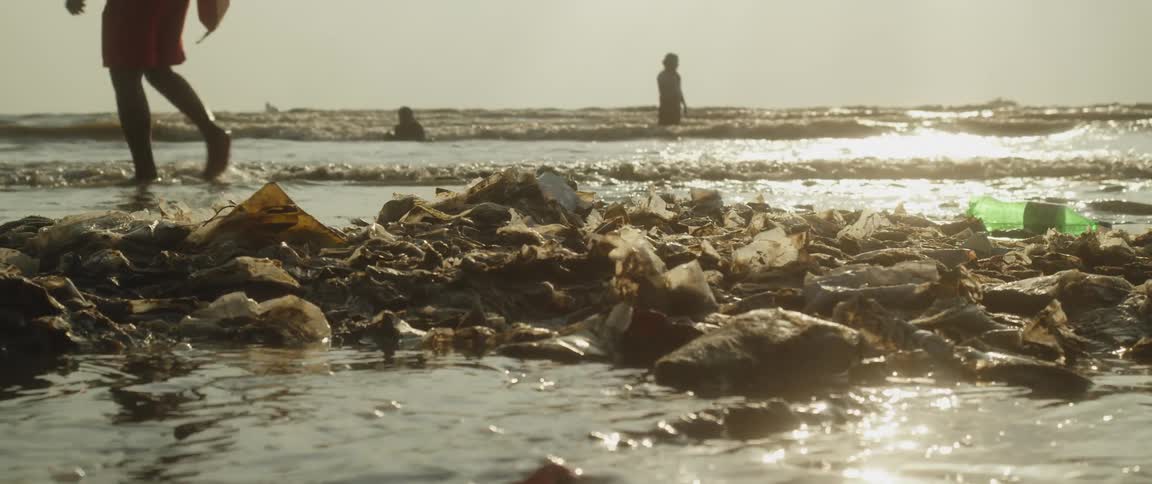 Garbage piled up on the beach as waves crash against it and people play in the water