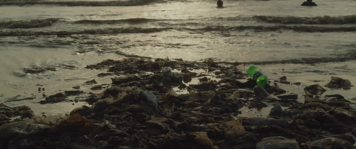 People are swimming at the beach as the sun shines through the clouds and waves roll into shore with plastic bottles and trash in the water