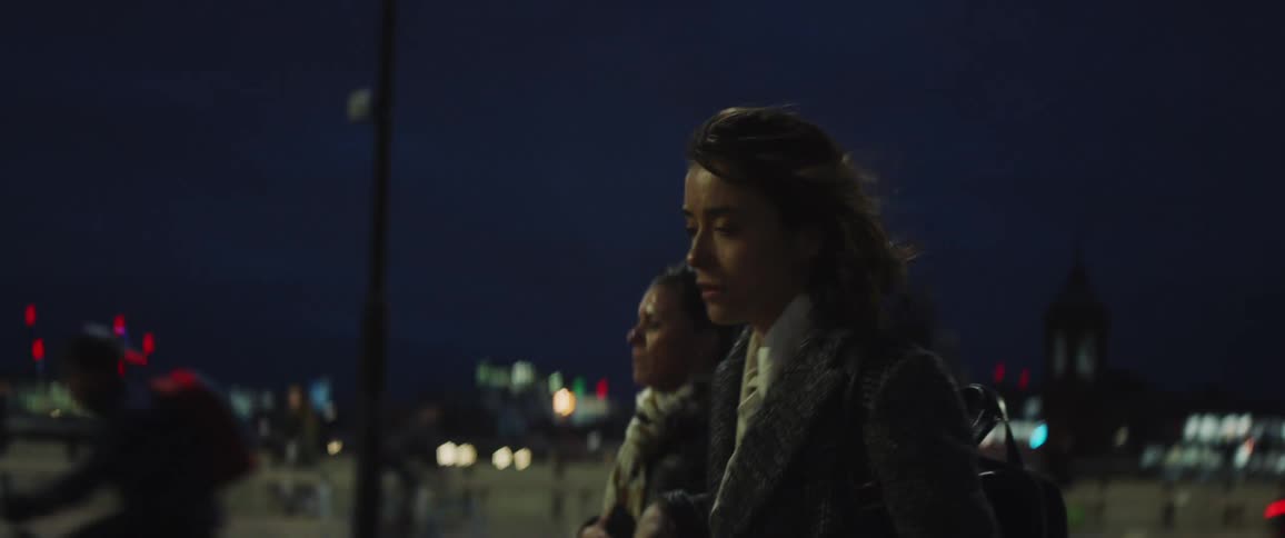 A woman walking down a sidewalk as traffic goes by on the street and other people walk by around her and she looks at her phone