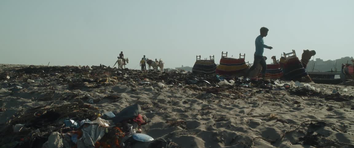 A boy walks past saddled camels in the sand looking at the trash and debris