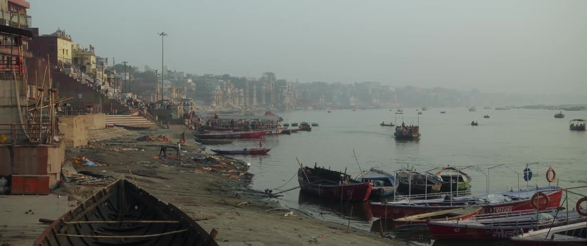 Boats are docked along the shore and the lake is filled with boats sailing and many people are working on the shore and walking near buildings along the coast