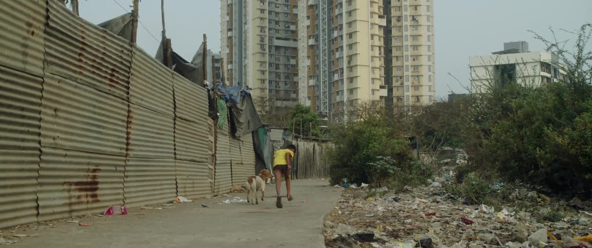 A little boy plays with a dog as they head down a slum street overlooked by a skyscraper