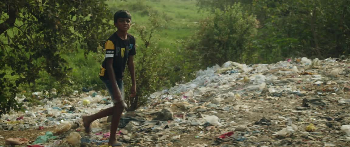 A young boy walking up a hill covered in garbage with green fields behind him