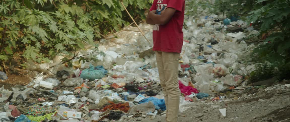 A teenage boy stands over a garbage dump in a forest looking sad