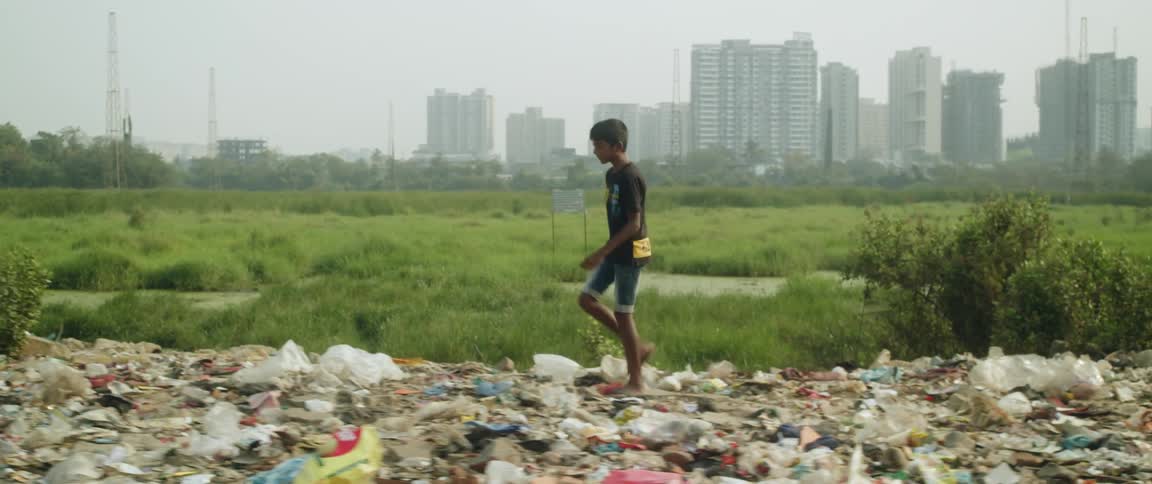 A young boy walking on a ridge covered in trash with tall city buildings behind him