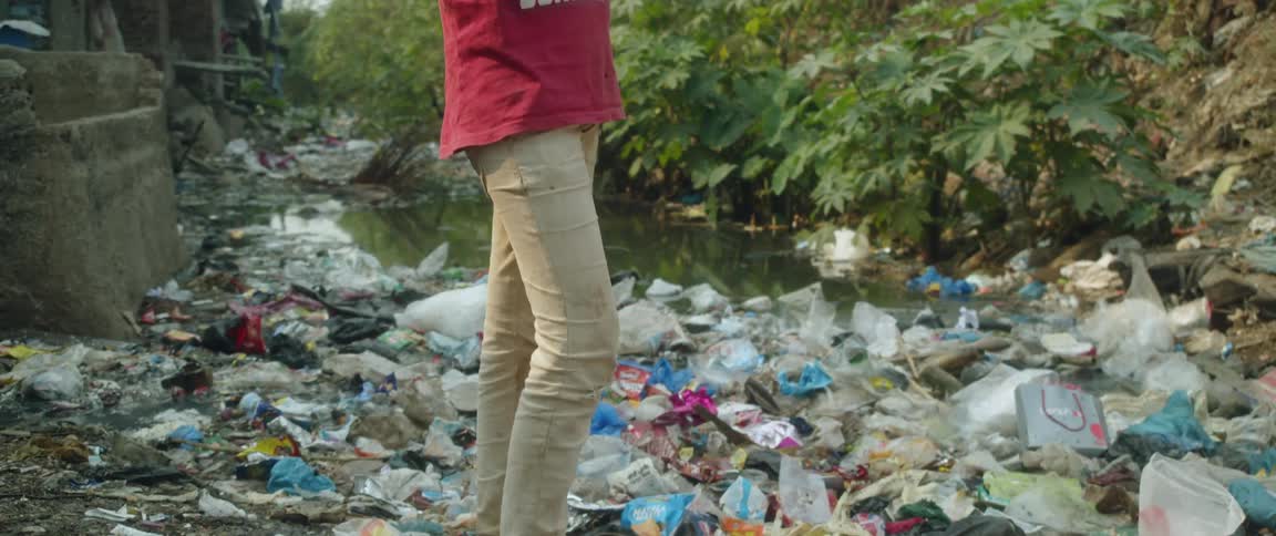A man stands on a riverbank surrounded by trash and litter