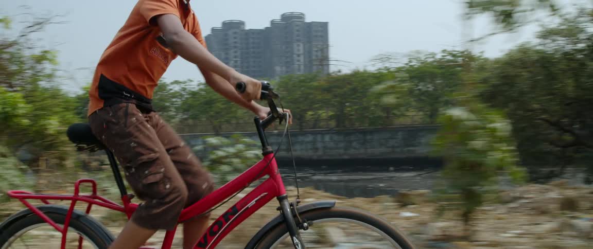 Young boy riding a red bicycle on a road near garbage and a city in the background