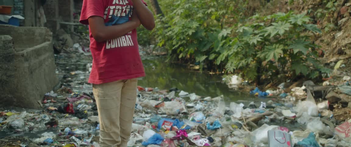 A young boy standing on a pile of garbage holding his hand over his noise while looking down around him