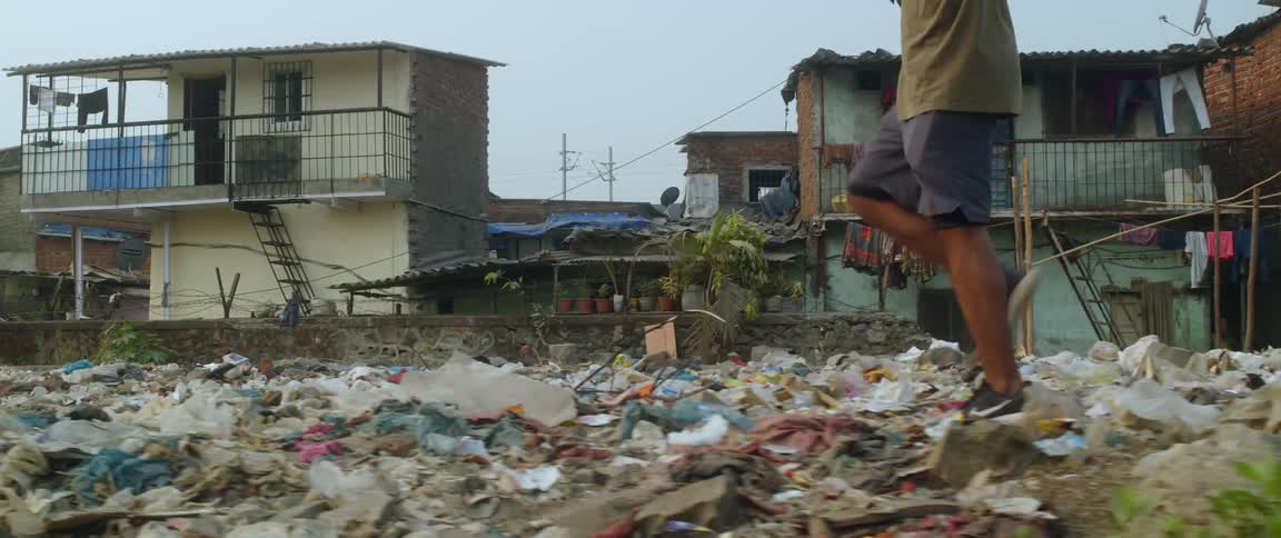 A boy is walking across garbage and trash outside of dilapidated and decrepit buildings in the slums