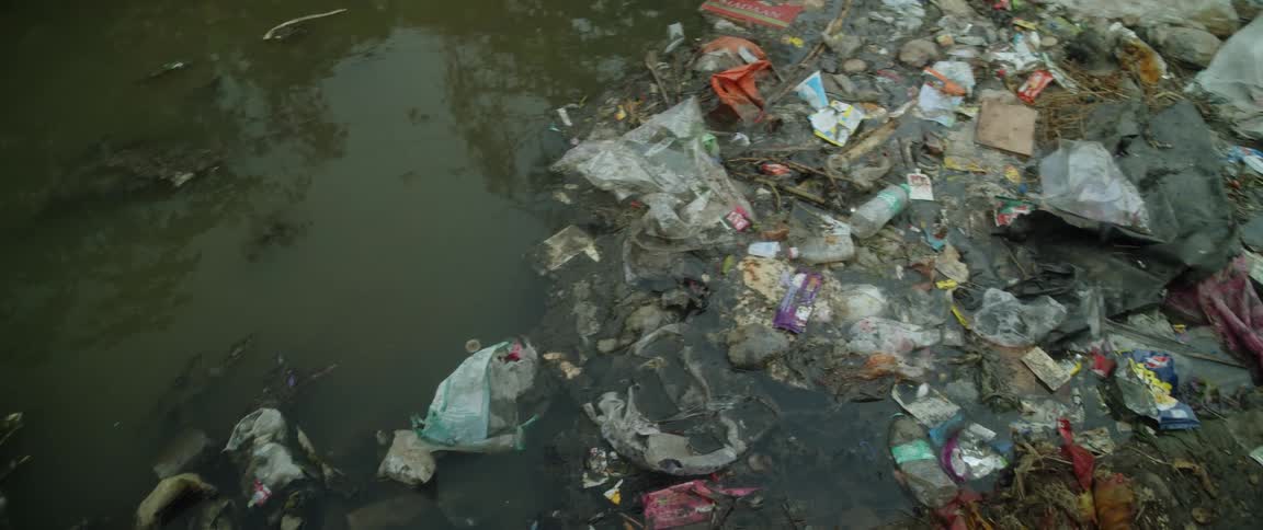 Garbage and debris are floating in a pond and trash covers a path as a bicycle leans against a wall