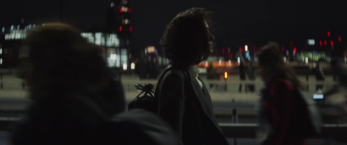 A woman walking down a sidewalk at night as traffic passes on the street and people walk by around her