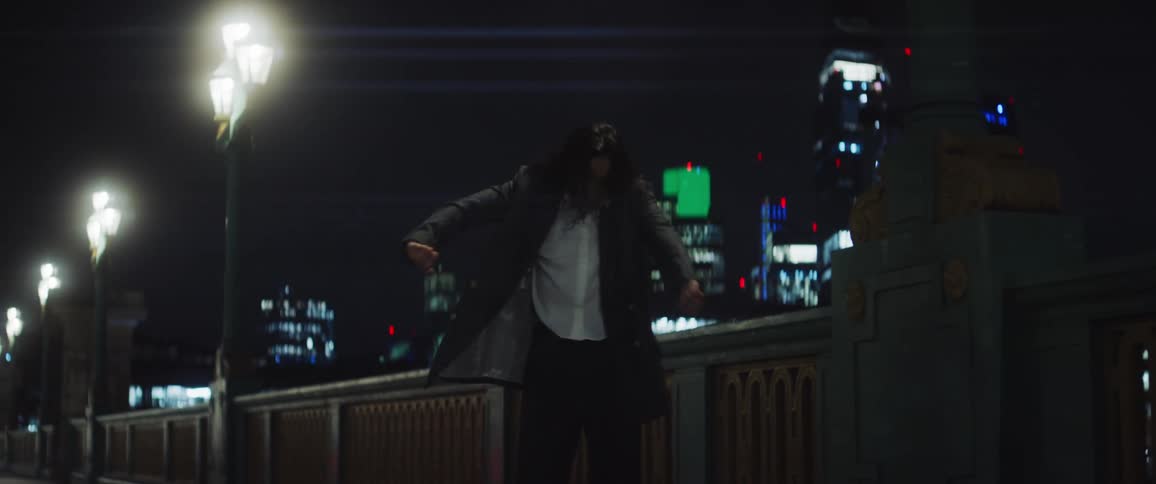 Woman dances near stone rail on bridge at night with city skyline in the background