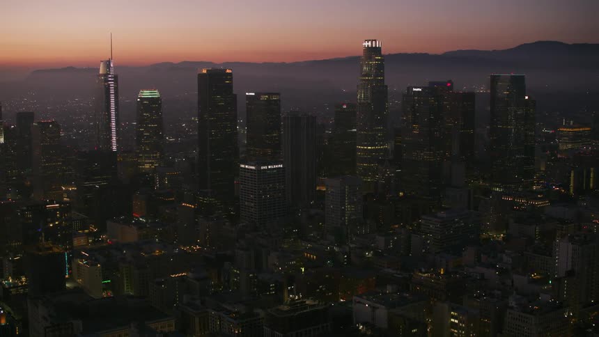 Towers and skyscrapers with modern architecture overlook a crowded city with lights on as the sun is setting and there are mountains in the distance
