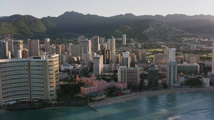 Waves roll into shore and over the beaches along the coast lined with tall buildings and towers of a big city with houses and buildings along the mountainside