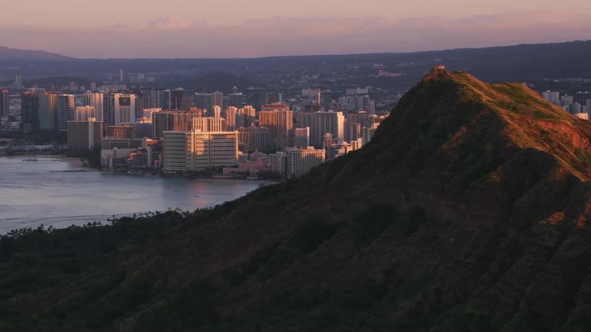 The sun shines on a mountain with buildings on the mountainside and a metropolitan city is between mountains along the coast as waves roll into shore