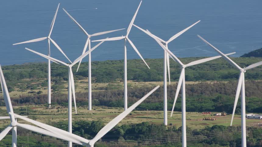 Wind turbines rotate in fields between woods near coast