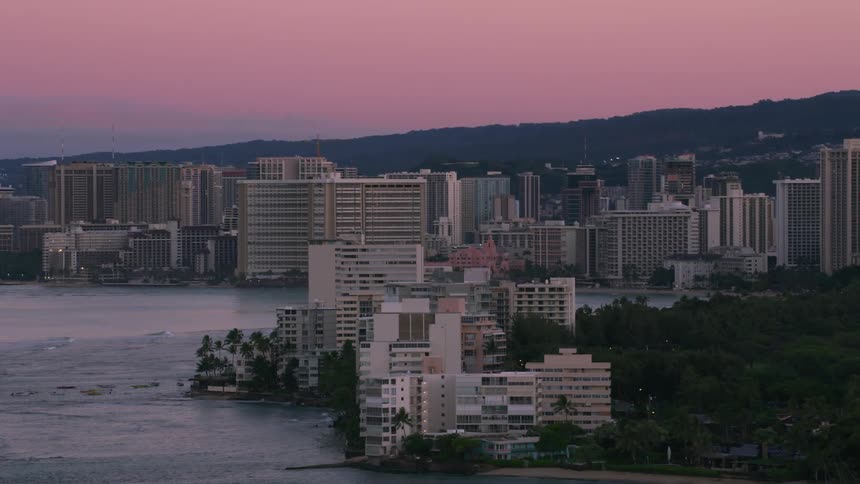 The sun shines on a mountain with buildings on the mountainside and a metropolitan city is between mountains along the coast as waves roll into shore