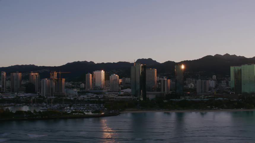 Sunlight shines on towers and tall buildings along the coast with boats in the marina and mountains in the distance