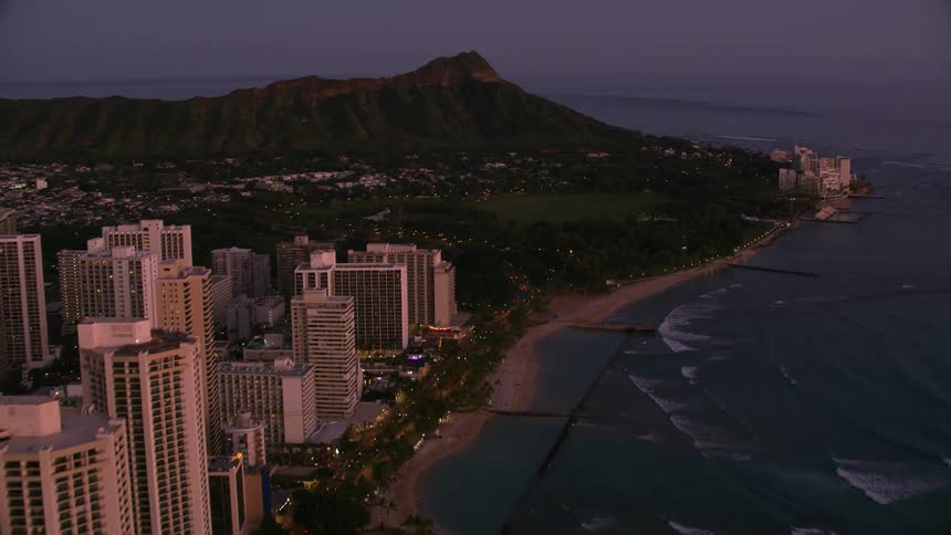 An aerial of a coastal city at dusk as waves crash against the coast with a mountain peak on the edge of the water