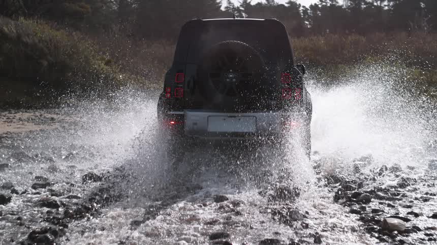 An suv drives through an almost dry and shallow river in a forest splashing water all around