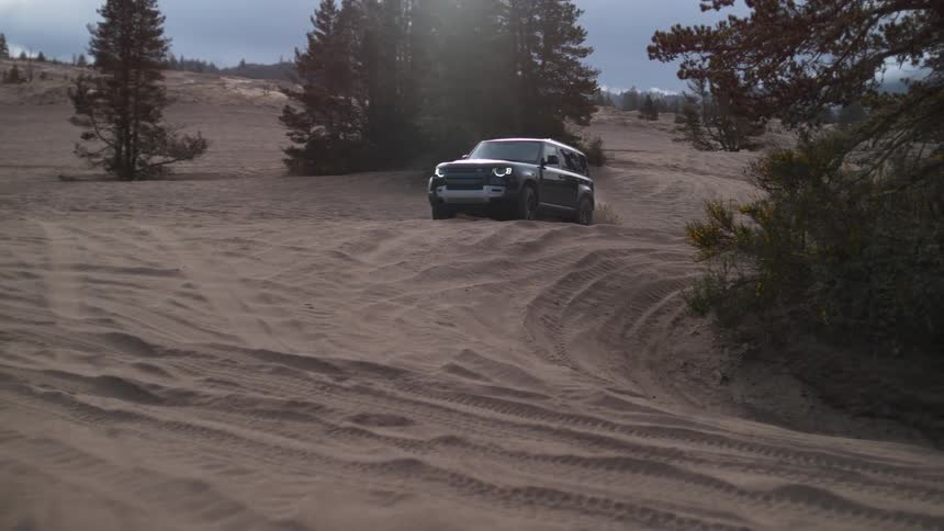 A black truck is traveling on a sand covered hill between trees under a cloudy sky