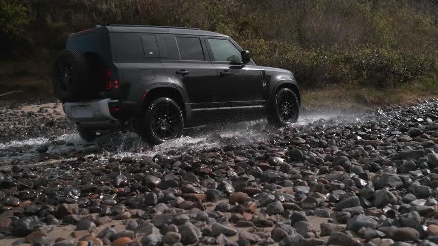 A black truck splashes as it travels through a water puddle on a stone road near where plants are growing on a hillside
