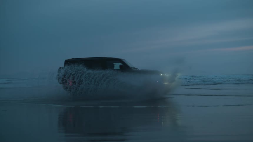 An suv drives through the water edge on a beach splashing water all around