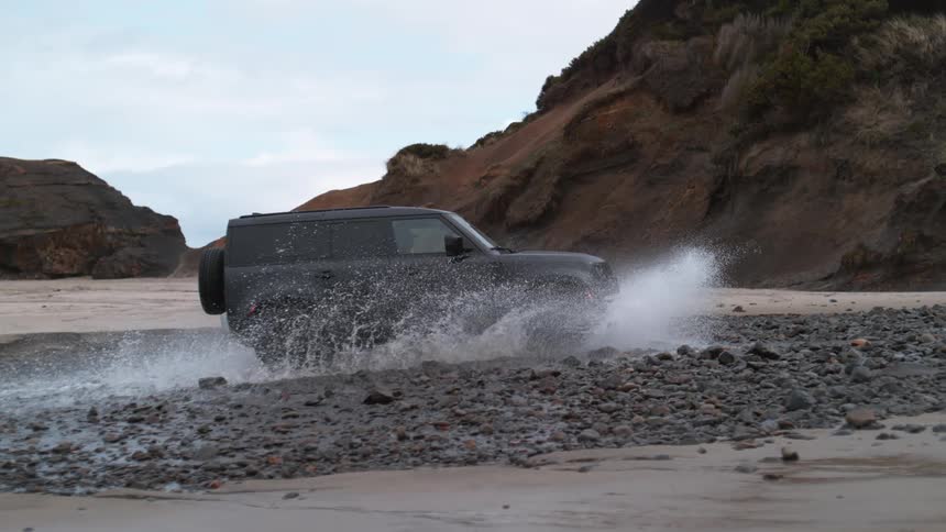 An suv drives through a puddle on a rocky beach splashing water around